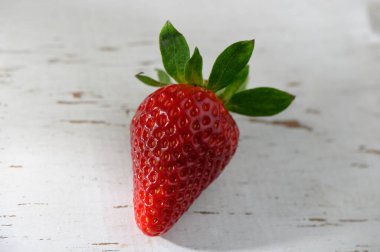 Canlı kırmızı bir çilek, yumuşak güneş ışığıyla aydınlatılmış yıpranmış ahşap bir masanın üzerinde duruyor. Meyve parlak yüzeyini ve yeşil yapraklarını sergiliyor, tazeliği ve yaz cömertliğini simgeliyor..