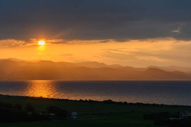 Güneş ufkun altına batarken, zengin altın renkleri sakin suları yansıtan gökyüzünü boyuyor. Uzak dağlar, dingin manzaraya derinlik katar. Saf bir sükunet anı yaratır..
