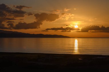 Gün sona ererken, parlak bir günbatımı ufku aydınlatıyor, sıcak renklerle gökyüzünü boyuyor. Yumuşak dalgalar kıyıya vurur ve suyun yanında huzurlu bir atmosfer yaratır..
