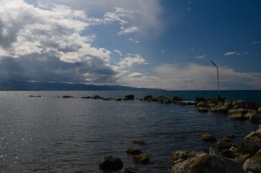 Sakin bir denizin kenarında, dalgalı bulutlarla dolu uçsuz bucaksız bir gökyüzünün altında pürüzsüz kayalar ortaya çıkar. Uzak dağlar çarpıcı bir zemin oluşturur, dingin atmosferi güçlendirir..