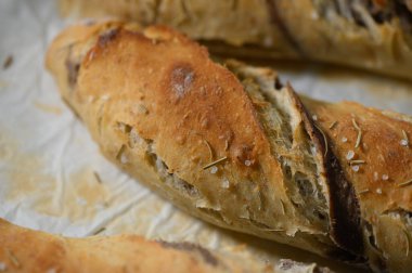 Altın kahverengi zanaatkar ekmek parşömen kaplı bir yüzeyde duruyor, deniz tuzu ve aromatik otlarla serpiştirilmiş çıtır bir kabuk sergiliyor, duyulara tat ve sıcaklık katıyor..