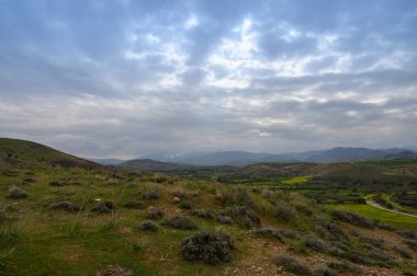 Huzurlu bir manzara, yumuşak alacakaranlıkla yıkanmış yuvarlanan tepelerle gözler önüne seriliyor. Yeşillik, dramatik bulutlarla dolu geniş bir gökyüzüne zıt düşer. Bu da doğanın ihtişamının sakin atmosferini canlandırır..