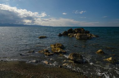 Dalgalar, su yüzeyindeki güneş ışığını yansıtan kayalık sahili ritmik olarak kucaklıyor. Görkemli bulutlar üzerimizde sürükleniyor, okyanus kenarında huzurlu ve ferahlatıcı bir atmosfer oluşturuyor..