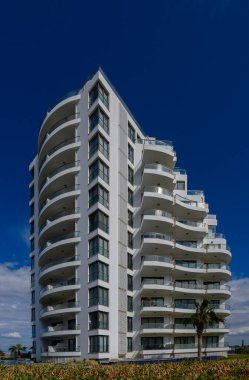 Çarpıcı bir modern bina, parlak kıvrımlar ve açık mavi gökyüzü ile çevrili geniş balkonlarla gururla yükseliyor. Tasarım, çağdaş kıyı yaşamını ve lüksü yansıtıyor..
