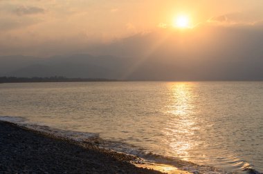 Güneş ufkun altına batarken, yumuşak altın ışınlar sakin sulardan yansıyarak çakıl taşlı bir sahili aydınlatıyor. Hafif bir esinti alacakaranlıkta huzurlu atmosferi karıştırır..