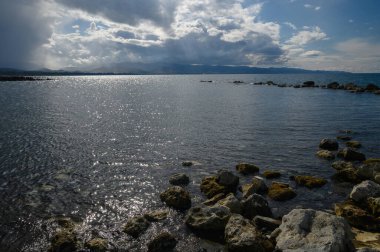 Dalgalar kayalar ve yosunlarla dağılmış engebeli bir kıyı şeridine nazikçe çarpar. Bulutlarla dolu dramatik bir gökyüzü değişen gölgeler oluşturur, günbatımında sakin bir atmosfer yaratır..
