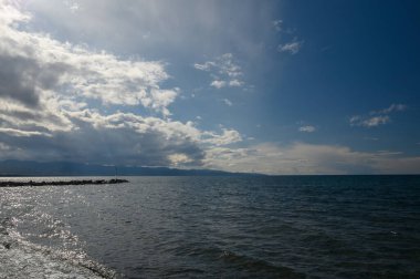 Dalgalar yavaşça kıyıya çarpıcı bir şekilde yaklaşırken bulutlar sakin suların üzerindeki alacakaranlık renklerini yansıtıyor. Huzurlu bir atmosfer kıyı manzarasını kaplar..