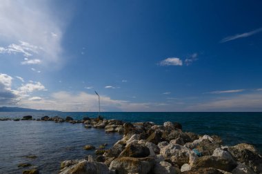 Kayalık oluşumlar engin mavi gökyüzünün altında parlayan okyanusa kadar uzanır. Yumuşak dalgalar taşlara çarpıyor bulutlar uzaklardan geçerken huzurlu bir atmosfer yaratıyor..