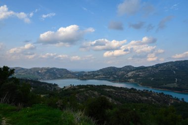 Parlak bulutlar berrak tepelerle çevrili sakin bir göl üzerinde canlı mavi bir gökyüzü boyunca sürükleniyor. Doğa, sıcak güneşin altında huzur içinde, dinlenmeyi ve tefekküre yol açar..