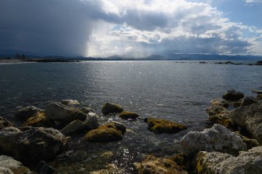 Karanlık bulutlar alacakaranlıkta uzak dağların üzerinde toplanırken dalgalar yosun kaplı kayalara nazikçe sarılır. Yansıtıcı su, değişen ışığın altında titreşiyor ve sakin bir atmosfer yaratıyor..
