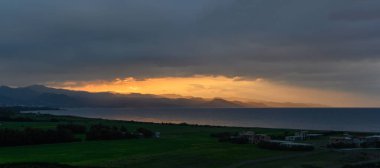 Güneş ufkun altına batarken, altın ışınlar engebeli kıyı dağlarını ve sakin denizi aydınlatıyor. Üzerimizde kara bulutlar sürükleniyor, akşam gökyüzünde dramatik bir kontrastlık yaratıyor..