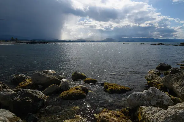 Karanlık bulutlar alacakaranlıkta uzak dağların üzerinde toplanırken dalgalar yosun kaplı kayalara nazikçe sarılır. Yansıtıcı su, değişen ışığın altında titreşiyor ve sakin bir atmosfer yaratıyor..
