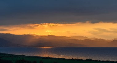 Güneş görkemli dağların ardında batarken altın renkler sakin okyanustan yansıyor. Dramatik bulutlar gökyüzüne derinlik katar, alacakaranlığın huzurlu güzelliğini arttırır..