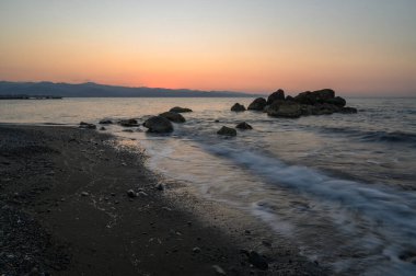 Güneş uzak dağların ardında batarken dalgalar yavaşça kayalık bir kıyıya vurur. Gökyüzü ve su boyunca yumuşak renkler bırakır. Huzurlu bir atmosfer manzarayı kaplıyor.