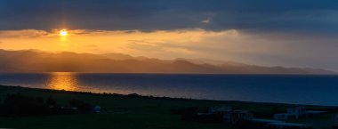 Altın ışınlar, turuncu ve mavinin renklerini yansıtan sakin sulara inerler. Aşağıda huzurlu bir köy dinlenirken, karanlık bulutlar ufku nazikçe kucaklıyor, alacakaranlıkta sakin bir atmosfer yaratıyor..