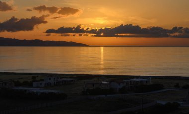 Canlı bir günbatımı, turuncu ve altın renkleri ile gökyüzünü boyar. Yumuşak dalgalar kıyıya vurur..