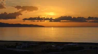 Güneş barışçıl bir okyanusun üzerinde batarken canlı renkler gökyüzünü boyuyor, suya altın yansımalar yapıyor ve dingin bir atmosfer yaratıyor. Uzak tepelerin siluetleri güzelliği arttırır.