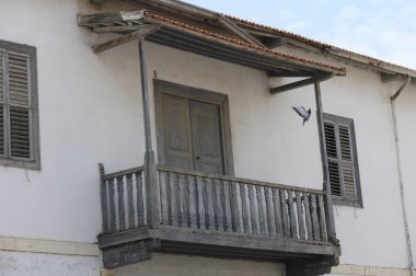 Yıpranmış ahşap bir balkon sergileniyor korkuluklar ve solmuş boyalar, dingin mavi gökyüzüne karşı uçan bir kuşun cesaretlendirmesi, özgürlük ve nostalji hissi uyandırıyor..