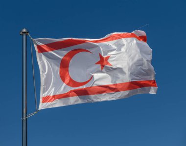 Güneş ışığıyla yıkanan Kuzey Kıbrıs bayrağı, kültürel gurur ve dayanıklılığı temsil eden hilal ve yıldız amblemini sergileyen parlak mavi gökyüzüne karşı güvenle dalgalanıyor..