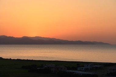 Güneş sakin suların üzerinde batarken, turuncu ve pembe çağlayanın yumuşak renkleri uzak dağların siluetlerini ve sakin bir kıyı şeridini aydınlatıyor. Sakin atmosfer yansımayı davet ediyor..