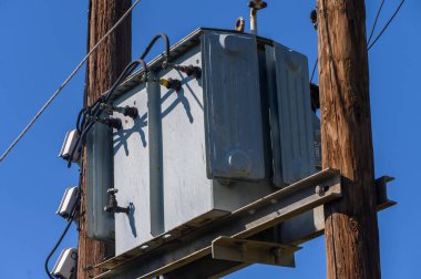 Bir güç transformatörü açık mavi gökyüzünün altındaki ahşap elektrik direğine güvenli bir şekilde bağlanarak banliyö ortamında elektrik altyapısının önemli rolünü vurguluyor..