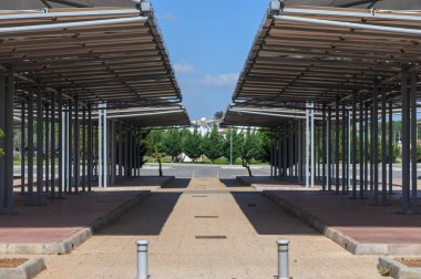 Tepe örtüsü yapıları açık bir alanda parlak güneş ışığıyla yıkanmış geometrik desenler oluşturur. Arkaplanda seyrek ağaçlar var, kentsel alana biraz doğa katıyor..