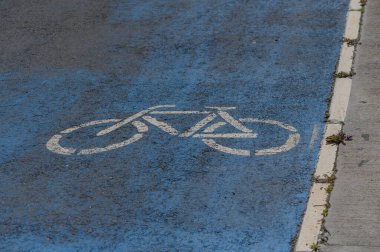 Canlı bir mavi bisiklet yolu işareti güneşli bir şehir caddesinde göze çarpıyor. Bu açık gösterge, hareketli şehirde bisiklet sürmeyi yeşil bir ulaşım tercihi olarak teşvik eder.