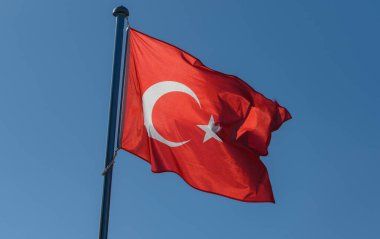 İkonik hilal ve yıldızıyla süslenmiş parlak bir Türk bayrağı, ulusal gurur ve birliği simgeleyen canlı mavi gökyüzünün altında enerjik bir şekilde dalgalanır..