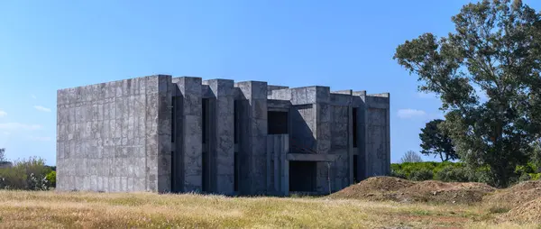 Çarpıcı bir beton yapı çimenli bir alanda modern tasarımı doğayla harmanlayarak yükselir. Uzun ağaçlar sahneyi çevreleyerek, açık gökyüzüne karşı ilgi çekici bir kontrastlık oluşturuyor..