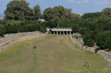 Parlak mavi bir gökyüzünün altında, canlı ağaçlar ve çimenli bölgelerle çerçevelenmiş, huzurlu bir nehir yatağı kuru yatar. Doğa gün ışığında bu huzurlu ortamda gelişir..