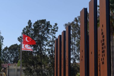 İki bayrak, ulusal kimlik ve kutlama duygusunu yansıtan uzun ve modern anıtlarla çevrili açık mavi gökyüzüne gururla dalgalanıyor. Huzurlu, davetkar bir atmosfer çevreyi sarar..