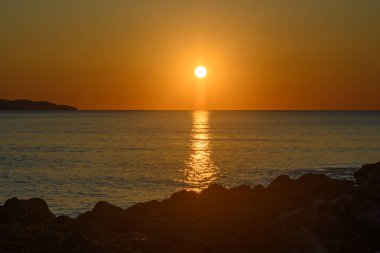 Nefes kesen bir günbatımı, sakin okyanus yüzeyinde parıldayan güneş ışınlarıyla ufku aydınlatır. Kayalık kıyı şeridi siluette duruyor, bu anın huzurlu atmosferini yükseltiyor..