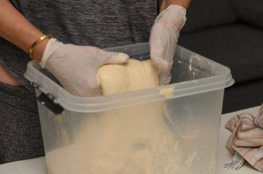 Bir çift eldivenli el, yumuşak bir hamuru ustalıkla plastik bir kapta yoğurur. Un yüzeyin tozunu alır, rahat bir mutfakta lezzetli bir ev yapımı ekmek oluşumuna işaret eder..