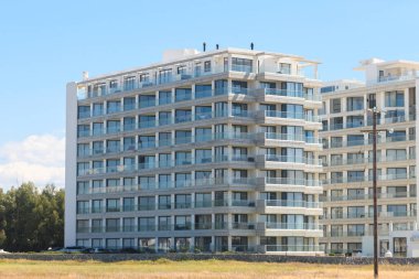 Gösterişli, çok katlı bir konut binasında açık mavi gökyüzünü yansıtan büyük cam pencereler bulunuyor. Yeşillik, modern tasarımla çelişiyor. Uyumlu bir şehir ortamı yaratıyor..