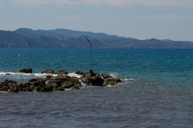 Dalgalar kayalık bir kıyı şeridine doğru süzülür, yalnız bir direk suyun kenarında bekçi olarak durur. Uzak dağlar yumuşak güneş ışığıyla yıkanır. Huzurlu ortamı güçlendirir..