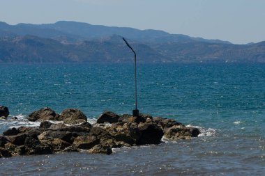 Dalgalar, güneşin ısıttığı kayaları hafifçe okşar. İnce bir direk, durgun bir kıyı bölgesini işaret ederken. Arka planda dağlar görkemli bir şekilde yükselir, barışçıl bir atmosfer yaratır..