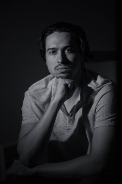 Bir adam düşünceli bir şekilde oturur, çenesini elinin üzerine koyar, gizemli bir duyguyu çağrıştıran derin gölgelerle çevrilidir..
