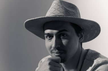 Hasır şapka giyen genç bir adam minimalist bir ortamda eli çenesinde dinlenirken düşünceli bir şekilde poz verir. Işık ve gölgenin etkileşimi onun özelliklerini vurguluyor, çarpıcı bir görsel yaratıyor..