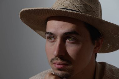 Çarpıcı yeşil gözleri olan genç bir adam eşsiz bir şapka takarken çenesini eline dayar. Yumuşak ışık, onun düşünceli ifadesini vurgular, yaratıcılık ve ilham atmosferi yaratır..