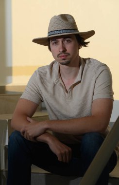 Hasır şapkalı genç bir adam merdivenlerde oturur, rahat bir yaz havası yayar. Sıcak güneş ışığı, öğleden sonranın tadını çıkarırken gevşemiş davranışlarını vurguluyor..