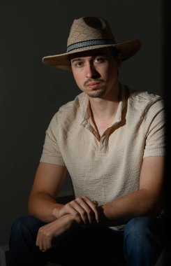 Düşünceli bir ifadeye sahip genç bir adam, hasır bir şapka ve açık renkli bir gömlek giyerek sessizce oturuyor. Loş ışık onun özelliklerini ön plana çıkarıyor, sanatsal bir atmosfer yaratıyor..