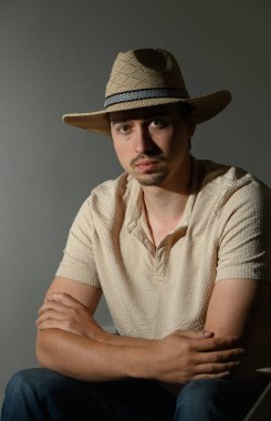 Hasır şapkalı genç bir adam sakince oturur, kollarını kavuşturur, hafifçe aydınlatılmış bir arka plana yaslar, sakin bir anda derin derin bir aura yayar..