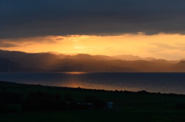 Güneş uzak dağların ardında batarken altın renkler göğü dolduruyor ve sakin sulara sıcak yansımalar yapıyor. Hava kararırken manzara doğanın güzelliğiyle parlar..