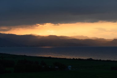 Güneş dağların ardında batarken nefes kesen bir manzara, denizler boyunca ılık altın bir ışık saçıyor ve bulutlarla kaplı gökyüzünü aydınlatıyor..
