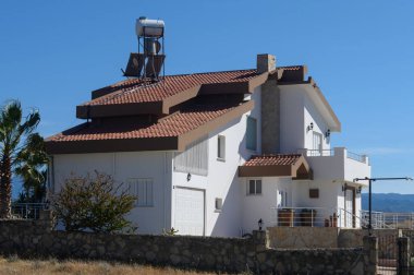 Şık bir villa tepenin yamacında duruyor, terracotta çatısı olan modern mimariyi sergiliyor. Bereketli yeşillik çevreyi sararken, güneş sakin manzaranın üzerinde parlıyor..