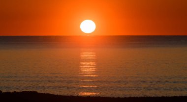 Gün sona ererken, güneş yavaşça alçalıyor, sakin denizde turuncu ve altının canlı tonlarını bırakıyor. Su yüzeyinde yansımalar parıldıyor, sakin bir atmosfer yaratıyor..