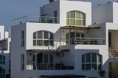 Günışığı, modern mimarinin parlak yüzeylerinde, berrak mavi gökyüzünün altında sakin bir kıyı atmosferiyle çevrili çok katlı ve balkonları sergiliyor..