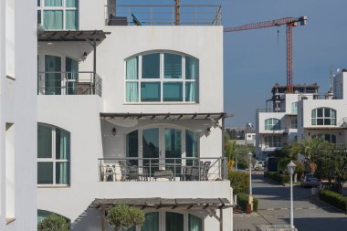 Mobilyalarla süslenmiş balkonlar, modern bir apartman kompleksinin ortasındaki iyi bakımlı bir yürüyüş yoluna bakıyor. Arka planda bir inşaat sahası beliriyor ve günün atmosferine canlılık katıyor..