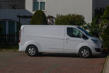 Gaziveren Kıbrıs Rum Kesimi - 16.04.2025 Sabah saatlerinde bir iskan bölgesine beyaz bir minibüs park edilmiştir. Ağaçları çevreleyen ve güneşli bir gökyüzü çevrede sakin ve sakin bir hava yaratıyor..