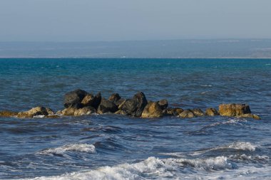 Parlak güneşin altında, sakin kıyı manzarası dalgaları zarif bir şekilde engebeli kayalarla etkileşime geçirir ve suyun kenarında sakin bir atmosfer yaratır..